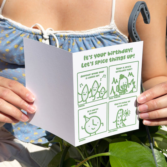 Person holding a card with humorous birthday message and illustrations, surrounded by green leaves.