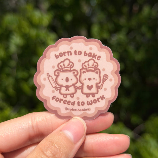 Sticker with cartoon characters saying born to bake, forced to work. Held by a hand against a blurred green background