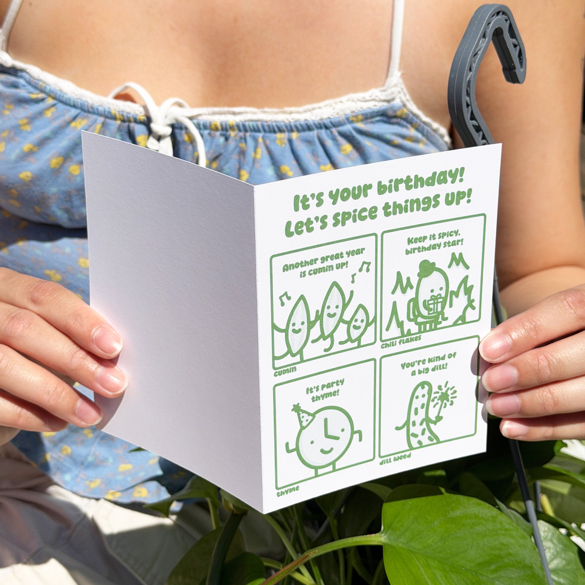 Person holding a card with humorous birthday message and illustrations, surrounded by green leaves.