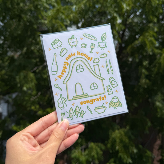 A hand holds a clear plastic sleeve containing a white card with green and yellow illustrations of a house, vegetables, and the words 'happy new home! congrats!' against a blurred green outdoor background.