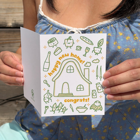 A person holding a white greeting card with green line drawings and orange text that reads 'happy new home! congrats!'. The person is wearing a blue floral dress, and the background shows a wooden deck.