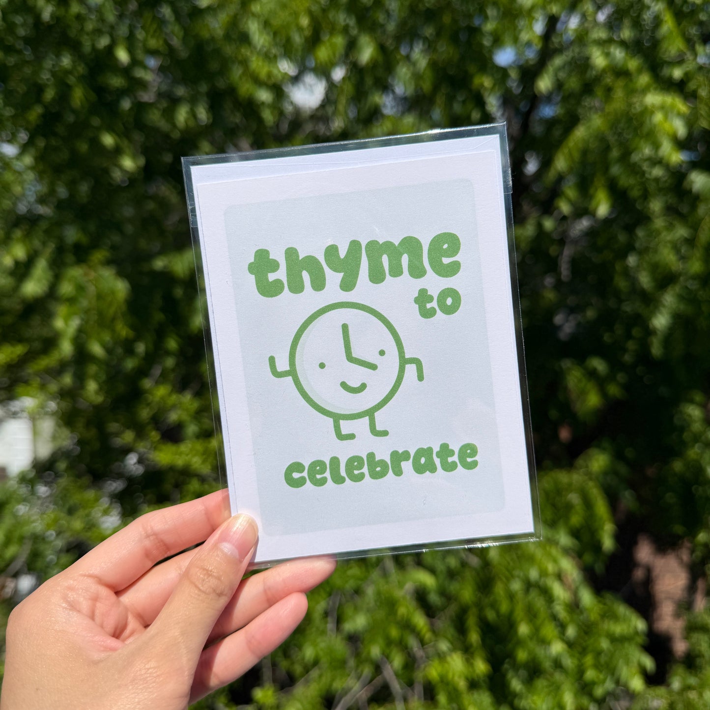 Hand holding a card with 'thyme to celebrate' text against a green leafy background