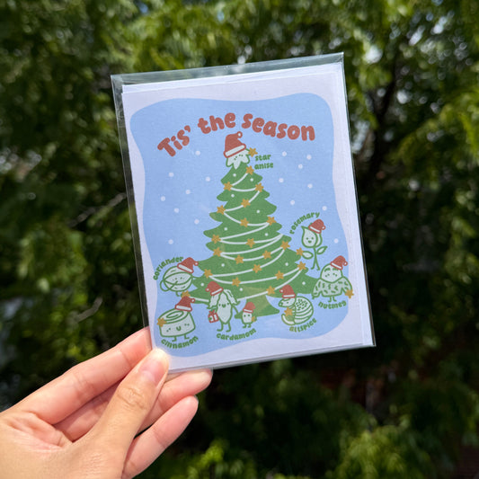 A hand holds a clear plastic sleeve containing a square card with a Christmas illustration and the text 'Tis' the season' against a blurred green outdoor background.