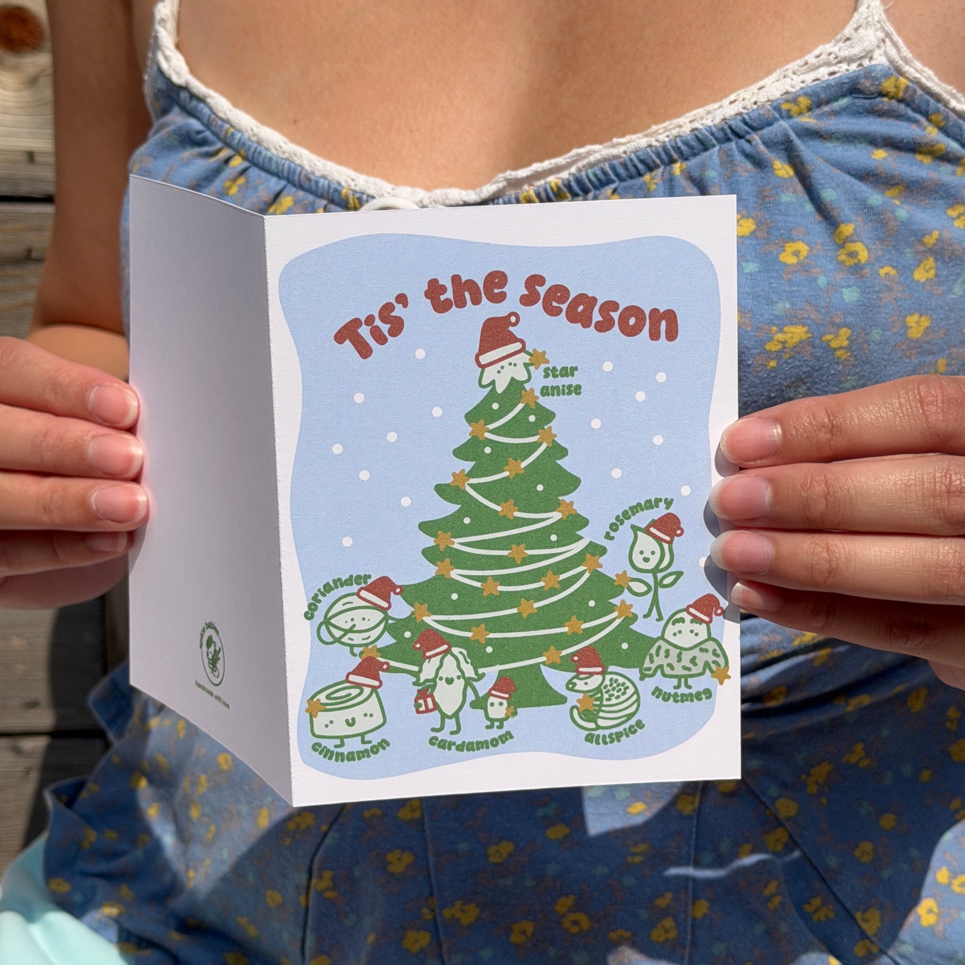A person holding a folded greeting card that reads "Tis' the season" with a whimsical illustration of a Christmas tree and holiday treats. The person is wearing a blue floral top, and the photo is taken outdoors in natural light.