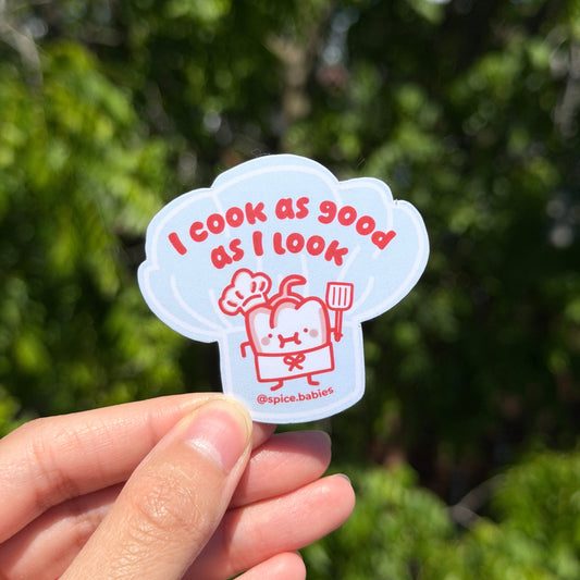 Hand holding a blue sticker with red text saying I cook as good as I look against a green blurred background