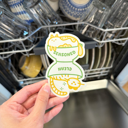 Hand holding a sticker with a cat design in front of an open dishwasher.