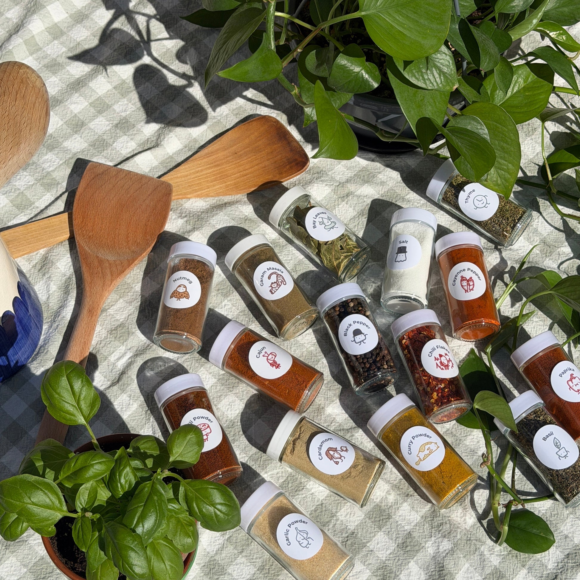 Spice bottles with labels on a checkered tablecloth with wooden spoons and plants.