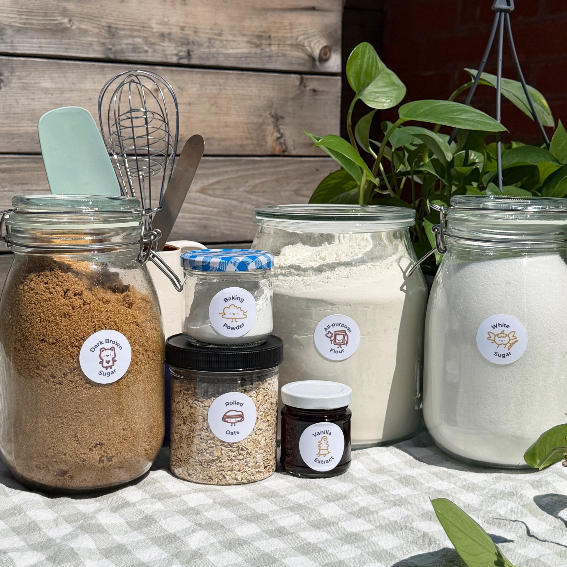 Jars of various baking ingredients on a checkered tablecloth with a wooden wall and plants in the background.