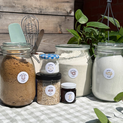 Jars of various baking ingredients on a checkered tablecloth with a wooden wall and plants in the background.