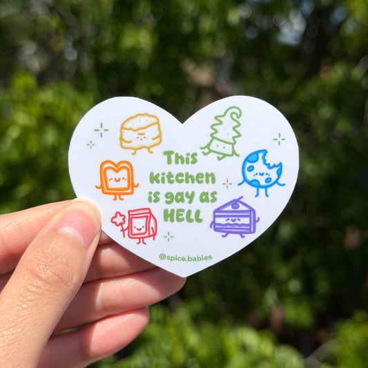 Heart-shaped sticker with colorful graphics that says this kitchen is gay as hell. Held by a hand against a green leafy background
