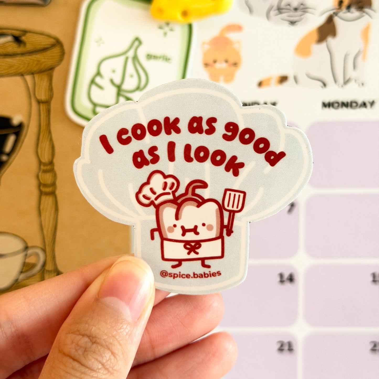 Sticker with 'I cook as good as I look' text held by a hand, with a calendar in the background