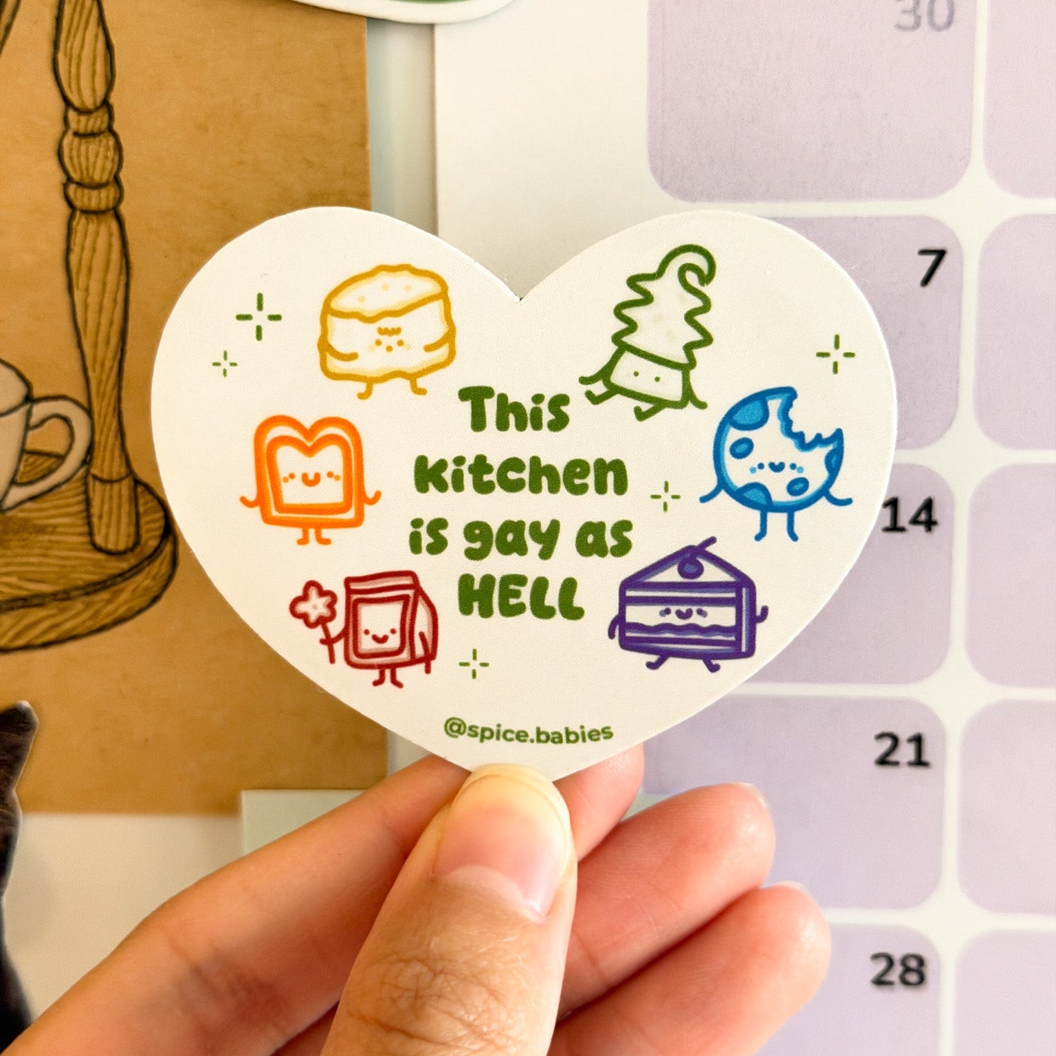 Heart-shaped magnet with colorful graphics saying this kitchen is gay as hell held over a calendar