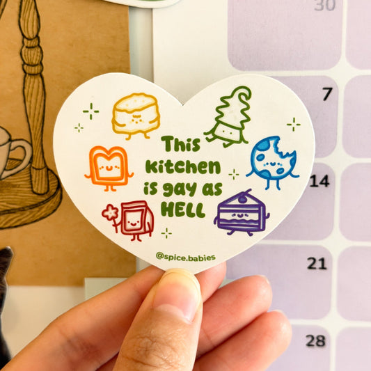 Heart-shaped magnet with colorful graphics saying this kitchen is gay as hell held over a calendar