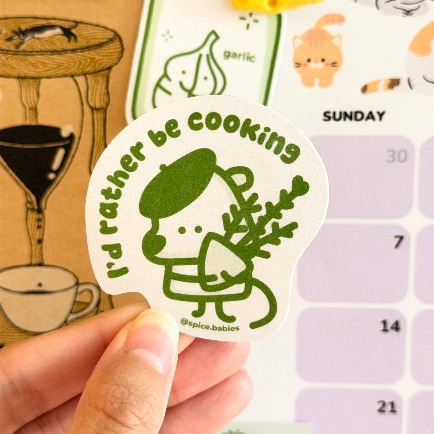 Hand holding a sticker with a character and text 'I'd rather be cooking' against a calendar background.