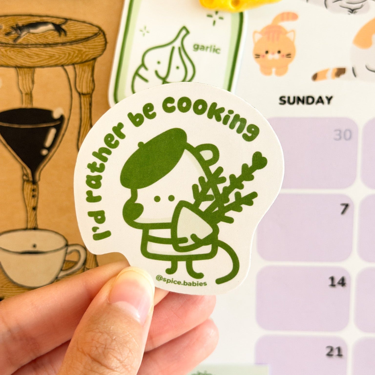 Hand holding a sticker with a character and text 'I'd rather be cooking' against a calendar background.