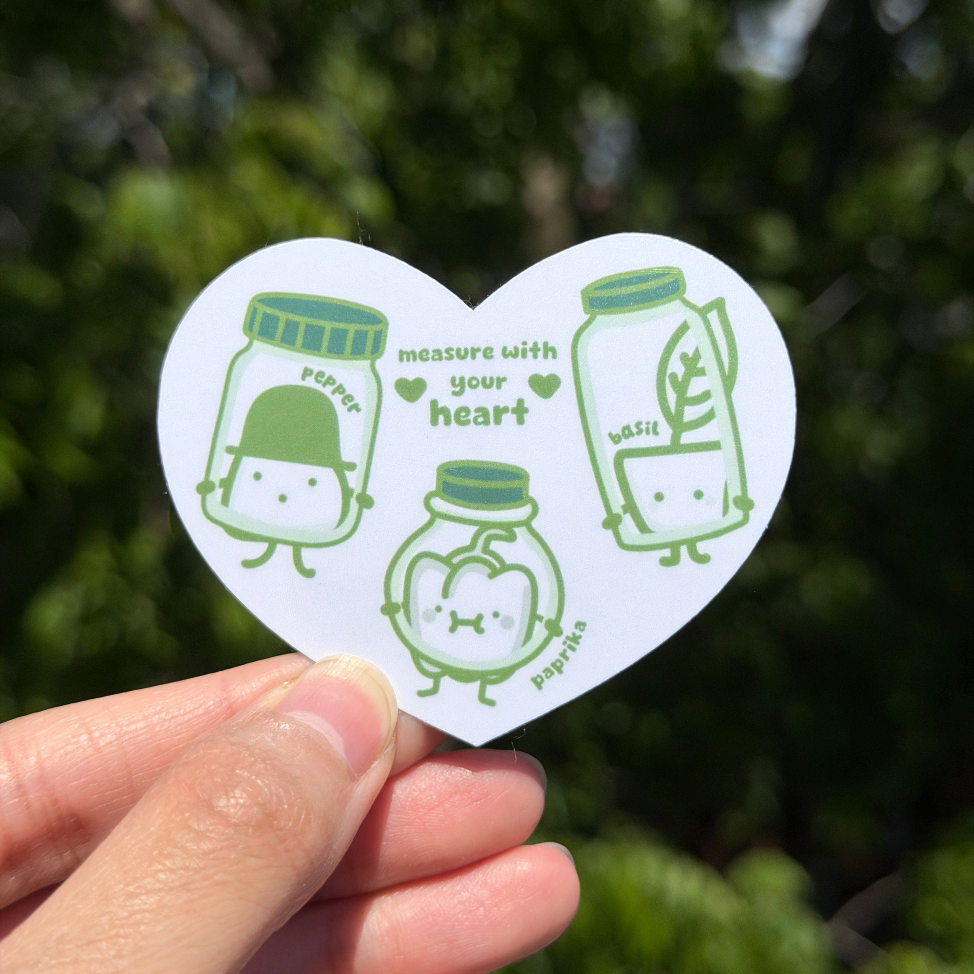 Heart-shaped sticker with green illustrations saying measure with your heart. It is held by a hand against a blurred green background