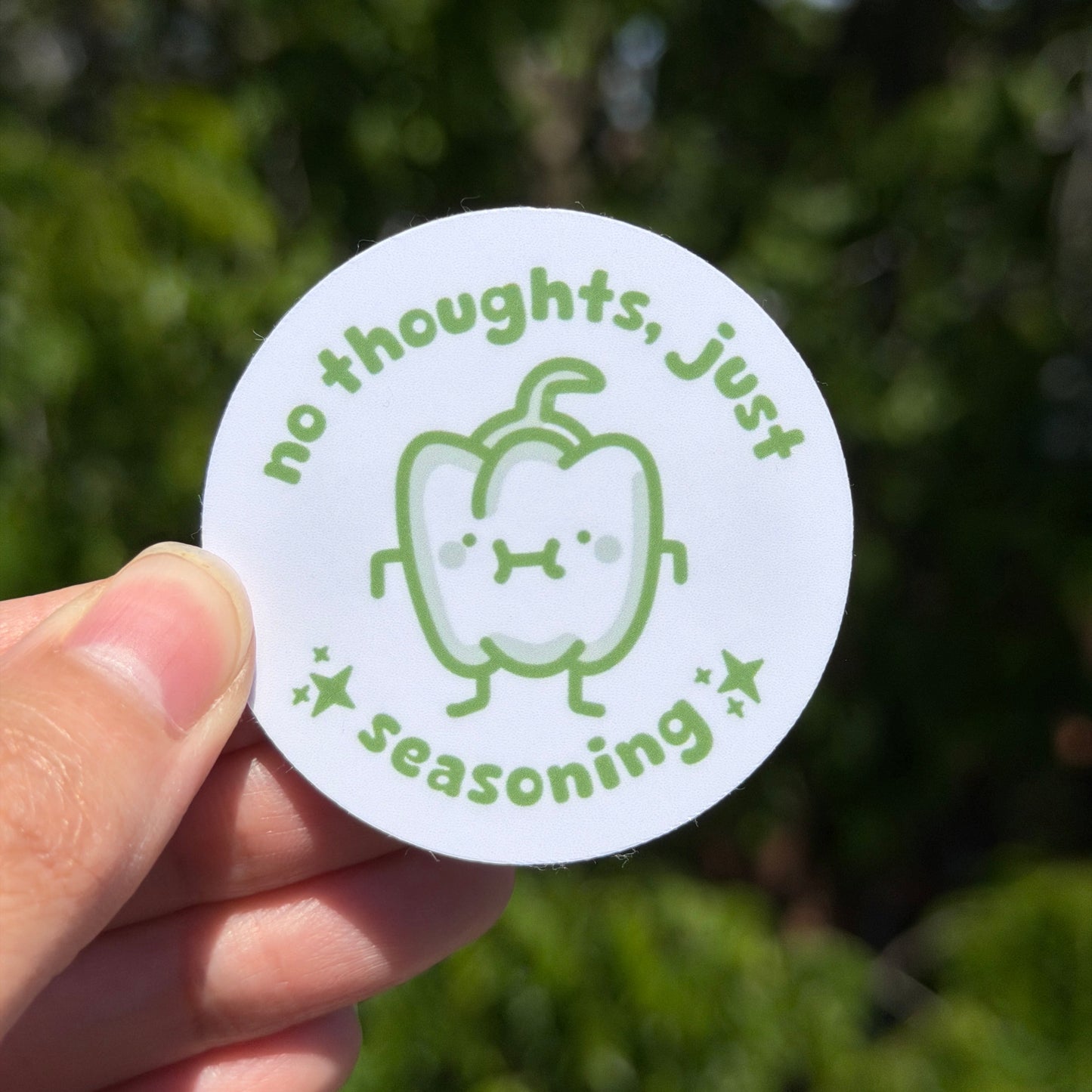 Circular sticker saying no thoughts just seasoning. Held by a hand against a blurred green background