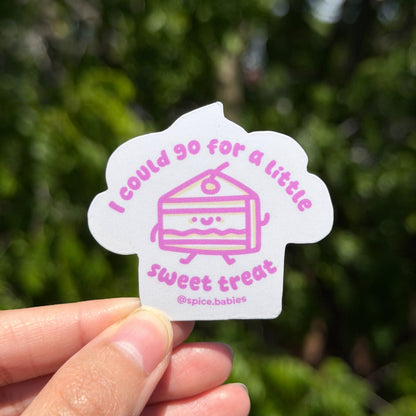 Hand holding a sticker saying I could go for a little sweet treat and a cake illustration against a blurred green background