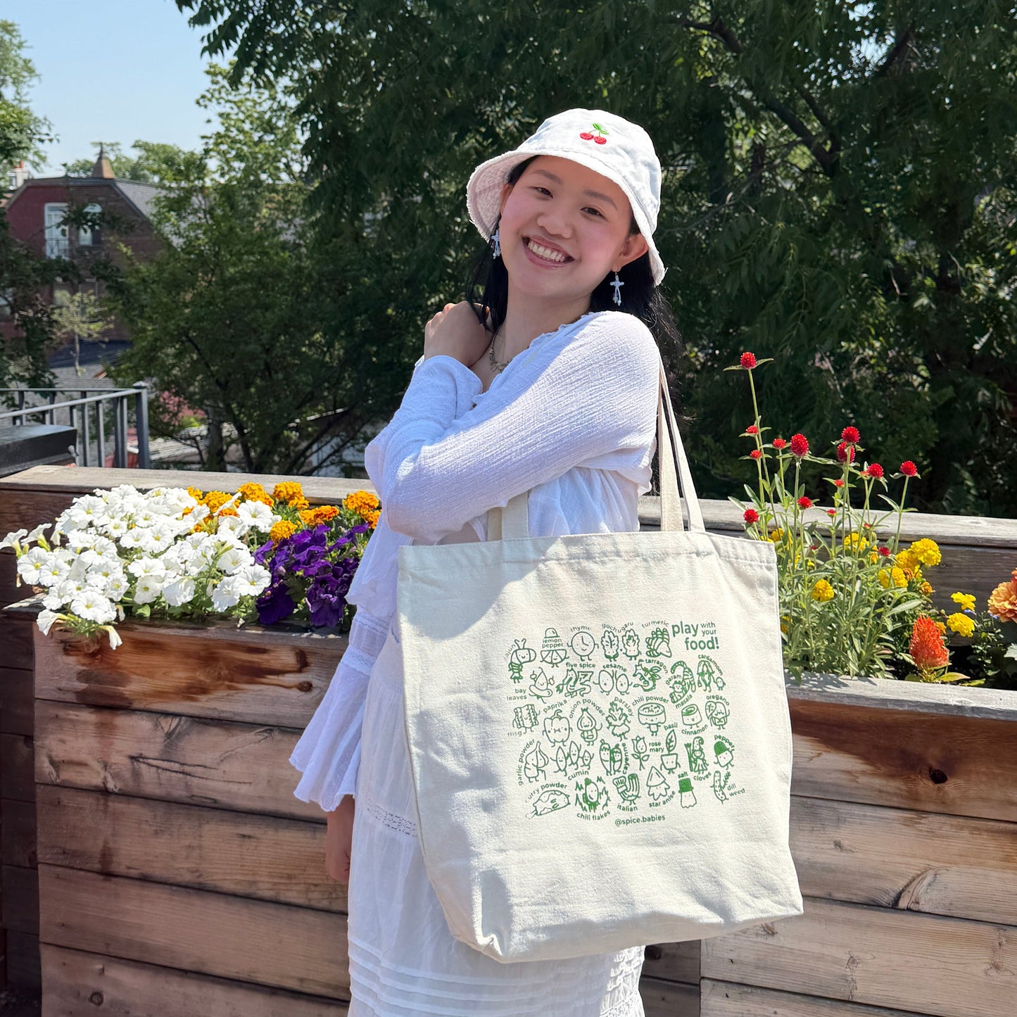 A woman wearing a white dress and a white bucket hat stands on a sunny outdoor balcony, holding a cream canvas tote bag with green text and illustrations. They are apart of the spice babies collection. The background features lush green trees, a wooden planter box with colorful flowers, and a brick wall.