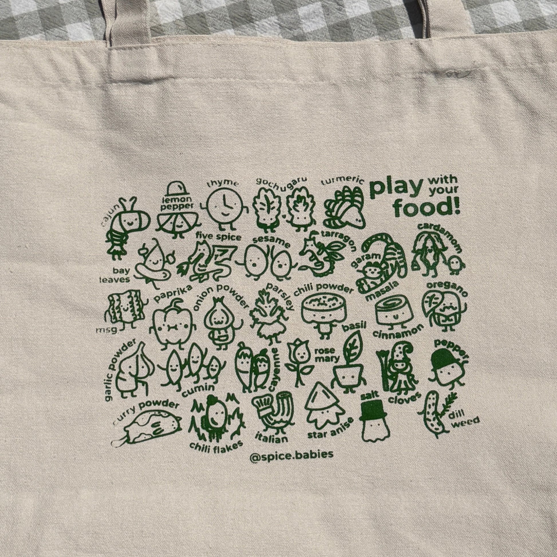 A natural canvas tote bag with green illustrations of spice babies and the text 'play with food!' printed on it, laid flat on a gray and white checkered blanket.