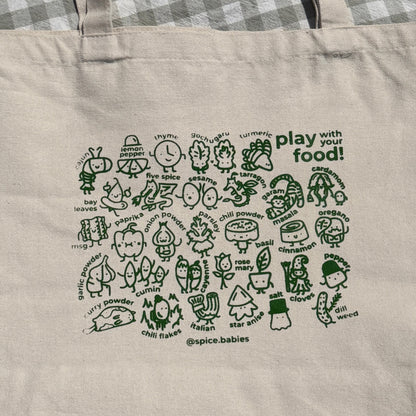 A natural canvas tote bag with green illustrations of spice babies and the text 'play with food!' printed on it, laid flat on a gray and white checkered blanket.